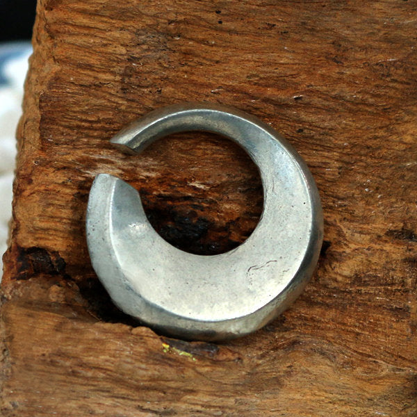 The HOLO Hoop Dangle Ear Weights in Silver by Kaya Movement, featuring a crescent moon-like design with an open gap, are beautifully displayed embedded in a rustic piece of wood. The ear weights appear to be handcrafted and contrast elegantly with the natural texture and color of the wood.